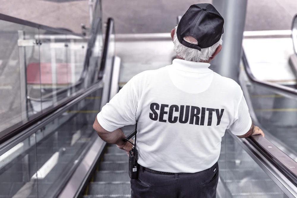 Servizi di vigilanza Guardia giurata di spalle in uniforme bianca con la scritta “Security”, mentre sorveglia dall’alto una scala mobile in un’area pubblica, pronta a intervenire per garantire la sicurezza.